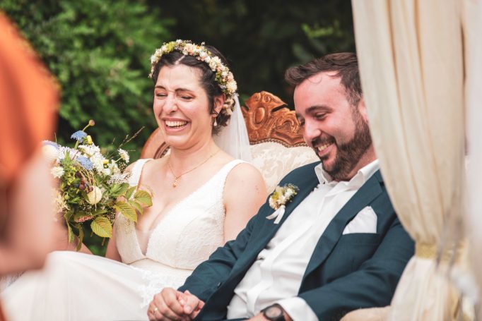 Braut und Bräutigam sitzen lächelnd auf einer Couch, von Blumen umgeben.